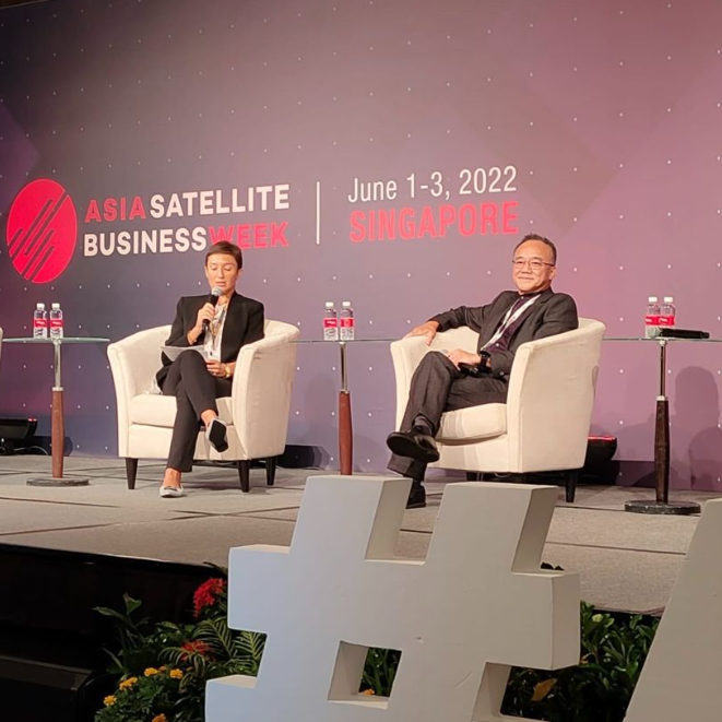 Two individuals sit in white armchairs with microphones on a stage at Asia Satellite Business Week in Singapore.